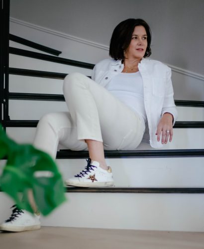 Midlife women wearing monochromatic look with ivory jeans, white tank top and white denim jacket and golden goose sneakers.