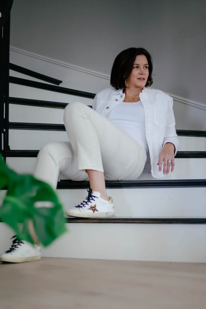 Midlife women wearing monochromatic look with ivory jeans, white tank top and white denim jacket and golden goose sneakers.