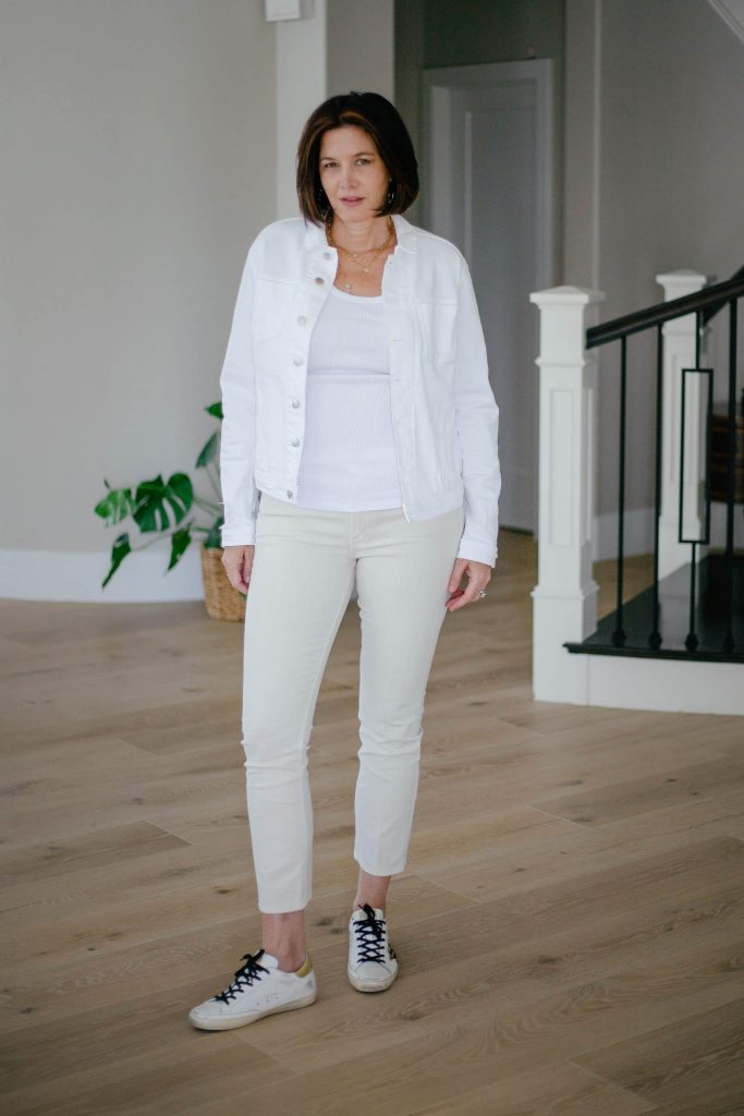 Midlife women wearing monochromatic look with ivory jeans, white tank top and white denim jacket and golden goose sneakers.