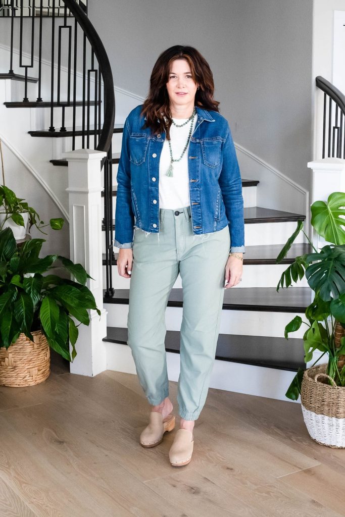Midlife women wearing clogs, pale green pants, cream puff sleeve shirt, jean jacket and necklace