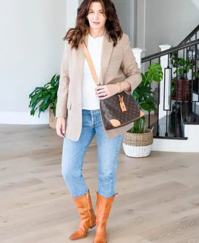 Midlife woman wearing cowboy boots, jeans, white tee and oversized blazer carrying Louis Vuitton purse.